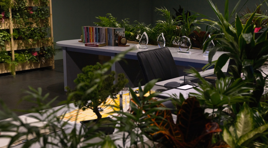'A Petropolis in a Garden with a Long View'. 2024–26. Video installation with laser-etched crystal, acrylic awards, books, globe-shaped bookholders, newspaper, magazine, mug, workspace accessories, office table and chair, wall shelves, planter boxes, assorted plants, wallpaper. Installation view, Diriyah Contemporary Art Biennale 2026: In Interludes and Transitions, Riyadh, Saudi Arabia, 2026. Courtesy of the artist